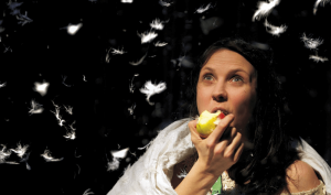 A woman eating an apple, as she watches white feathers fall down around her.