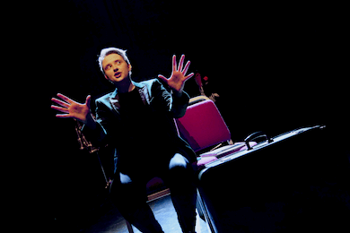 A person sitting on a chair, with a suitcase to their side, holding their hands up and looking away from the camera