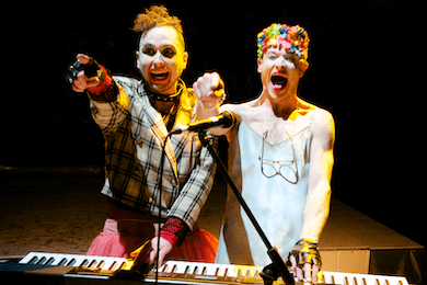 Two people standing at a keyboard, laughing and pointing out towards the camera.