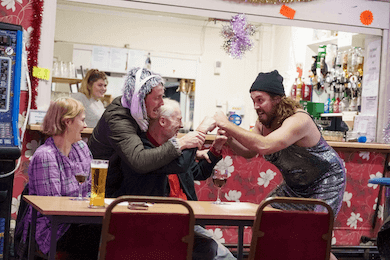 Two men in costume pretending to play fight with another man sat down, while two women look on laughing.