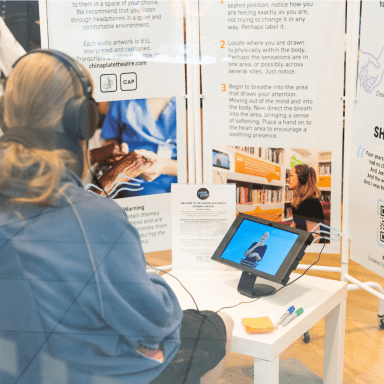 A person sat with their back to the camera, wearing a pair of headphones, whilst watching and listening to an audio piece being BSL interpreted on an iPad screen. In the background are several panels, displaying information and text about Humans Not Heroes. These panels act as a screen for a mobile listening station.