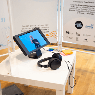 A close up of an iPad screen with a woman BSL interpreting an audio piece. Next to the screen is a pair of headphones, both of which are situated on a small white table. There are some post it notes and pens too.