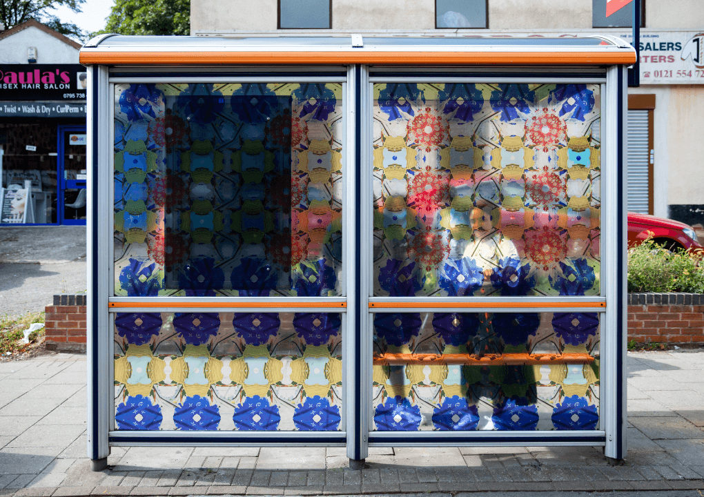 A bus stop decorated with a repeated patterned vinyl. A young woman can be seen through the pattern standing at the bus stop.