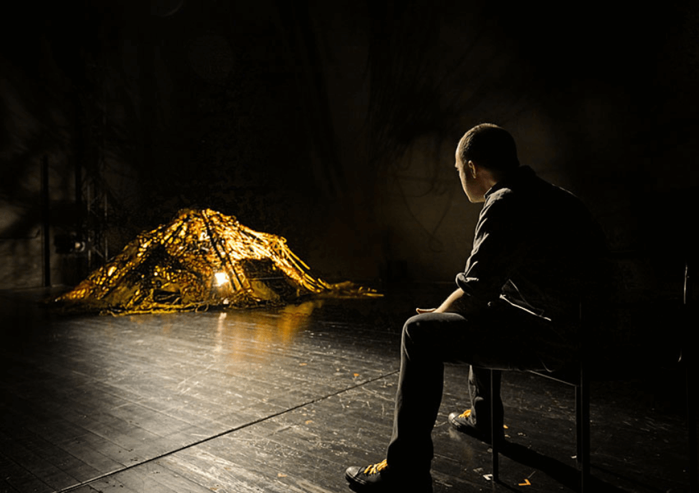 A pile of yellow wires is lite up in the left corner of the photo on a darkened stage. A man with his back to the camera is sitting on a chair looking at the pile of wires.