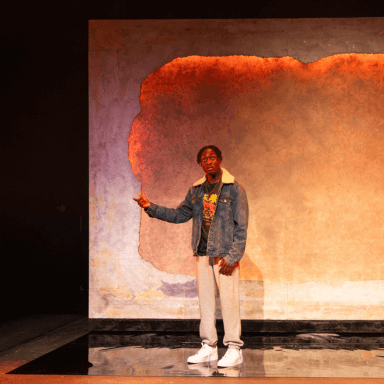 A young black man stood in front of a bright orange backdrop. He is wearing light grey joggers, white trainers and a denim jacket. His right arm is raised and pointing off to the side.