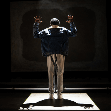 A young black man stood with his back to the audience, in a white square spotlight on stage. His arms are raised above his head.