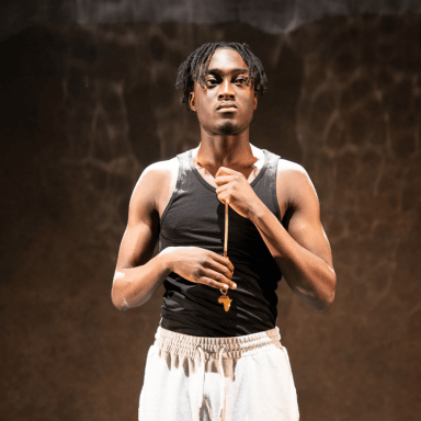 A young black man stood facing the audience, in a white spotlight on stage. He is wearing light grey joggers, a black vest and gold necklace.