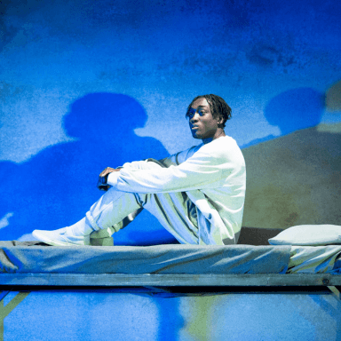 A close up of young black man sat on top of a bunk bed, with his knees tucked up. He is wearing a light grey tracksuit. The panel backdrop behind him is illuminated blue.