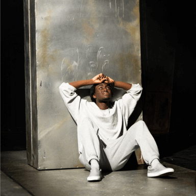 A young black man sat on the floor with his hands above his head. He is leaning against a large silver cabinet.