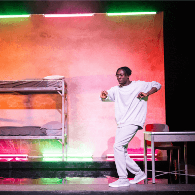 A young black man dancing on stage, with his arms raised. He is wearing a light grey tracksuit. Behind him is a backdrop and a bunk bed illuminated in pink and green hues.