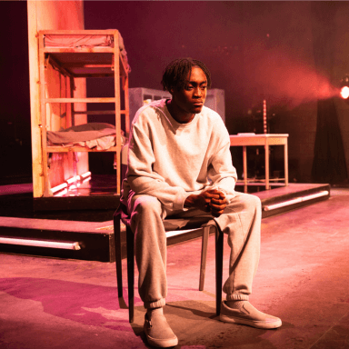 A young black man sat on a chair, resting his arms on his legs. He is wearing a light grey tracksuit. In the background is a bunk bed and table upon a raised platform. Spotlights are projecting pink and yellows hues.