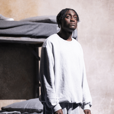 A close up of young black man looking upwards under a bright white spotlight. He is wearing a light grey tracksuit.