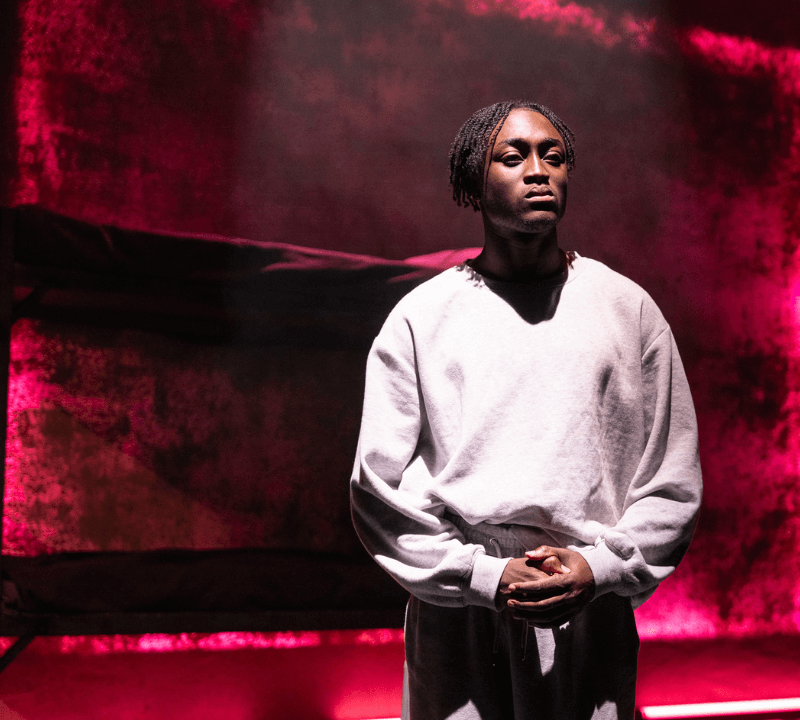 A young black man stood under a spotlight, on darkened stage. He is wearing a light grey tracksuit and has braided hair. A vibrant pink hue is shining through the backdrop of the set, which sits behind a bunk bed.