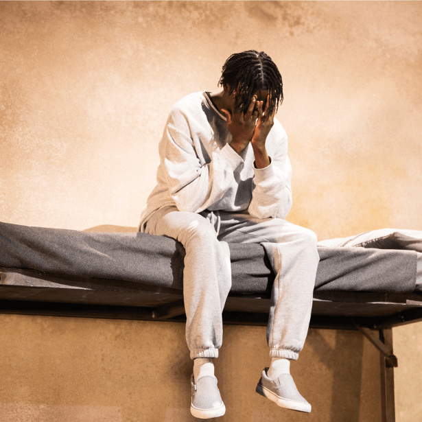 A young black man sat on top of a bunk bed, with his face in his hands.