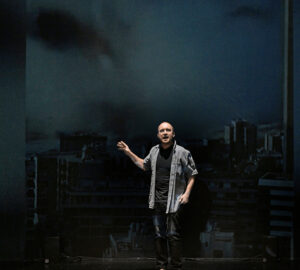 A man stands with his right arm raised slightly. He is on stage looking out towards the audience. Behind him is a video projection.