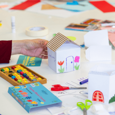 A view of a craft table, with various pens, pencils, crayons and scissors. In the centre is a paper house which someone has created and drawn on. On the side it says the word 'sanctuary'.