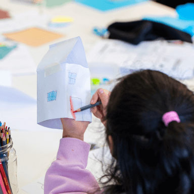 A young child, with their back to the camera, colouring in a paper house they have made.