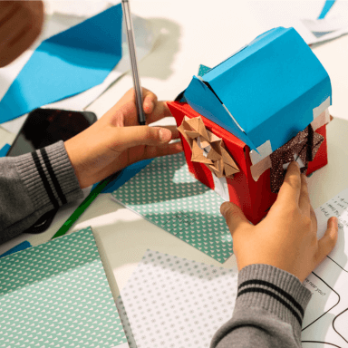 A close up of a young child's hands and the paper house they have created. It has a blue roof, red sides and origami folded windows.