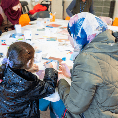 A woman and a young child sat at a table doing some colouring in and crafts together. They both have their backs to the camera.