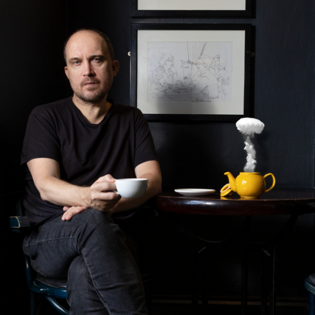 A man sitting at a coffee table, holding a mug, with a yellow teapot on the table with smoke escaping.