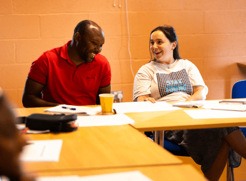 Two people sitting side by side at a desk, laughing and smiling.