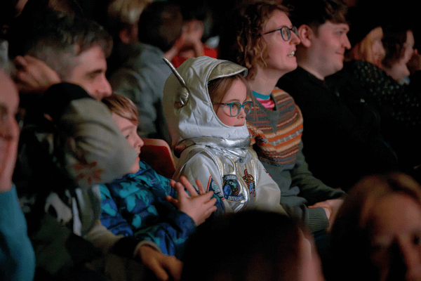 A young girl is sat in the audience, dressed as an astronaut, looking towards the stage out of shot.
