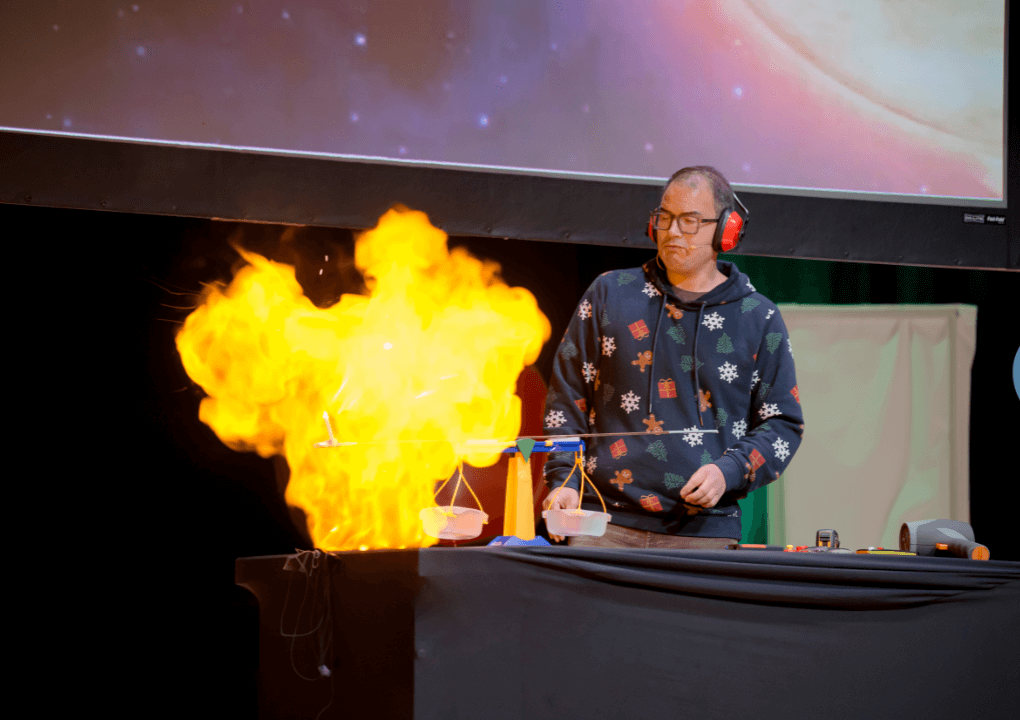 A man wearing a Christmas jumper and red ear defenders and glasses, is stood behind a table, as a large flame of fire escapes from a balloon which has been ignited by a candle at the end of some scales.