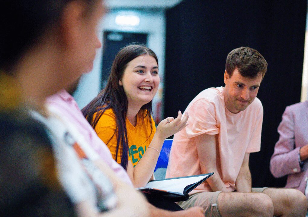 A young woman with long brown hair and purple highlights appears to be facilitating a group discussion. She has a book resting on her lap and a pen in her hand.