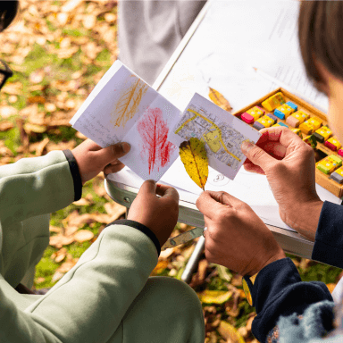 A close up of two people holding a paper book, map and leaf in their hands. They appear to be sitting at a table in a park.