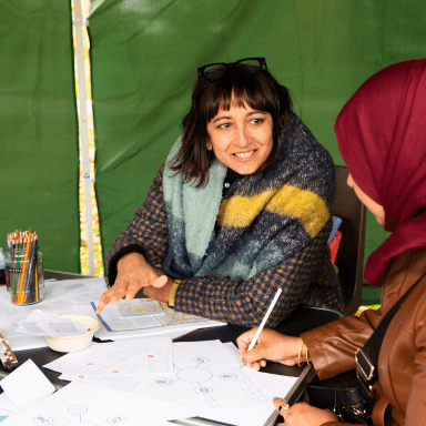 A woman is looking towards a person sat them to her. They appear to be in conversation. The woman is wearing a big scarf over a coat and has a pair of glasses resting on her head.
