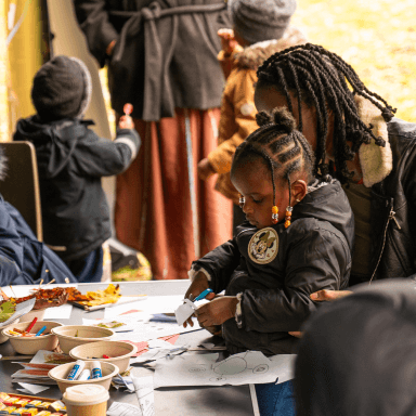 A young child cutting out some paper whilst sat on an adults lap. There is a table with crafty items in front of them.