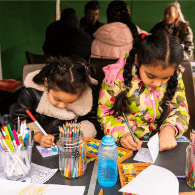 Two young children sat side by side at a table colouring in some small folded paper books.