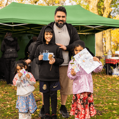 A man is stood with three children in a park. The three children are holding up the arts and crafts they have been making.
