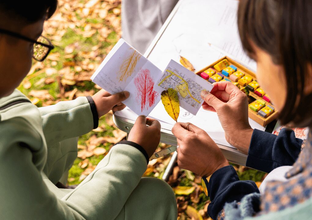 A close up of two people holding a paper book, map and leaf in their hands. They appear to be sitting at a table in a park.