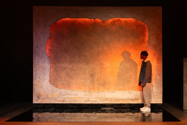 A young black man stood side on in front of a bright orange backdrop. His silhouette can be seen against the backdrop. He is wearing light grey joggers, white trainers and a denim jacket.