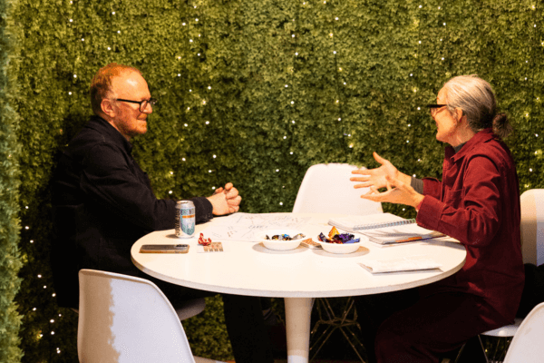 Two people are sat at a round table having a discussion. The table is situated in a pod like space which has fake greenery around it and fairy lights.