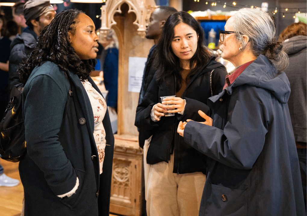 Three people stood talking to one another. One of three is holding a drink in their hands.