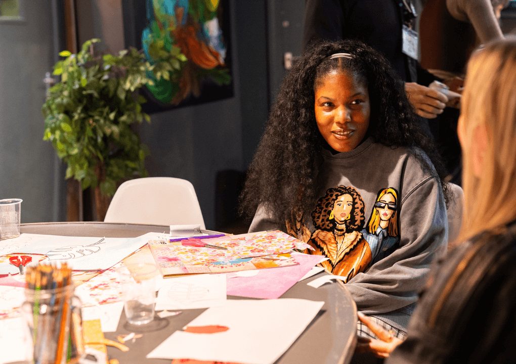 A person with long curly dark hair is sat facing the camera. They are sat at a table talking to another person, whose back is to the camera.