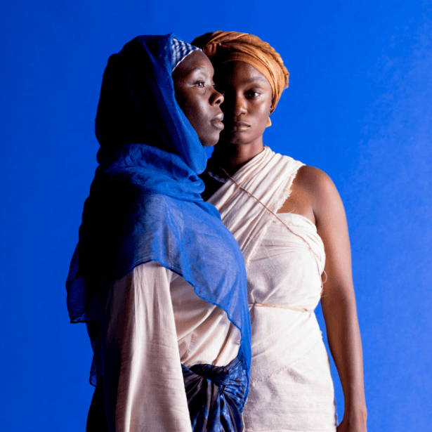 Two women are stood against a dark blue background. One woman is facing directly towards the camera. The other woman is stood sideways slightly overlapping the other woman.