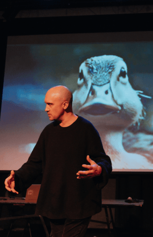 A man stood in front of a large projection screen with a duck on it. He is wearing a black jumper and gesturing with his hands out in front of him.
