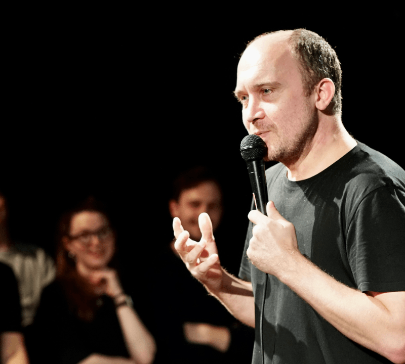 A photograph of a man holding a microphone to his mouth. Some audience members can be seen in the background.