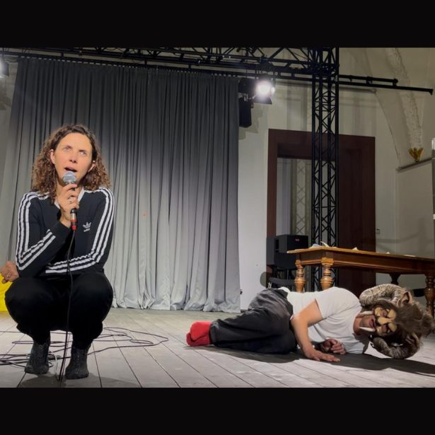 Two people performing, one is crouched down holding a microphone, the other is lying on the floor wearing a mask.