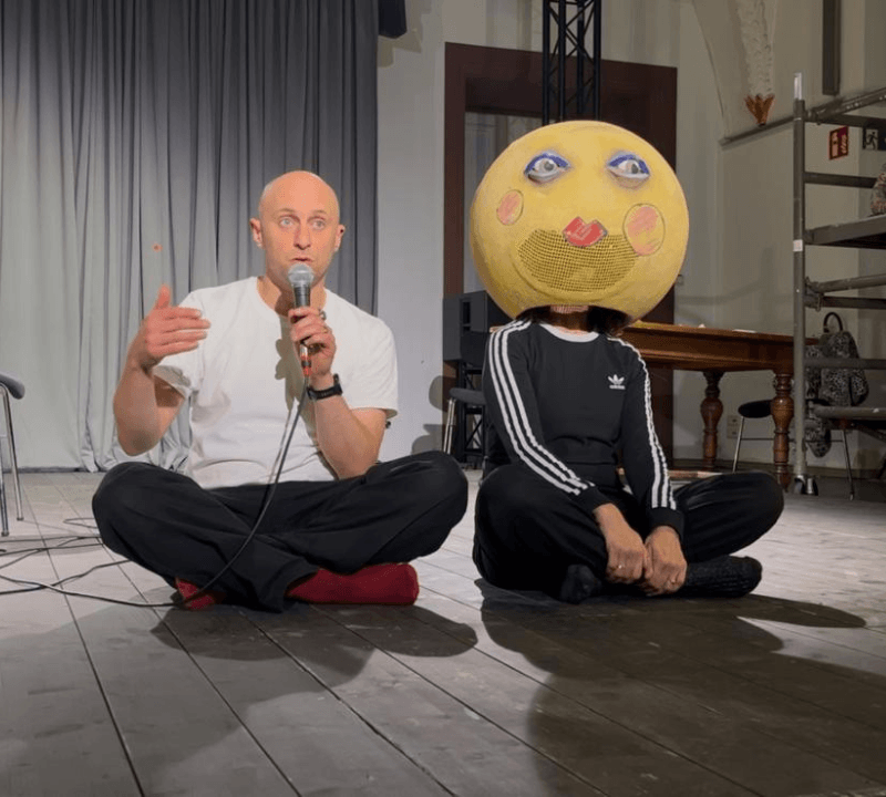 Two people sat on the floor crossed legged. One of them is speaking into a microphone, the other is wearing a large yellow ball on their head which has a face drawn on it.