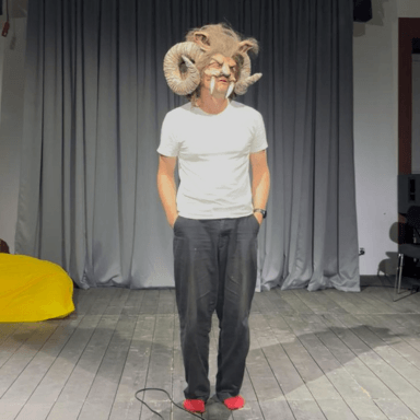 A man stood with his hands in his pockets, wearing a mask which has horns, tusks, ears and hair.