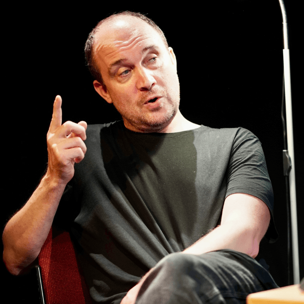 A man sits at a table, with a laptop in front of him. Behind him is a small standing lamp. He is looking out and talking towards the audience.