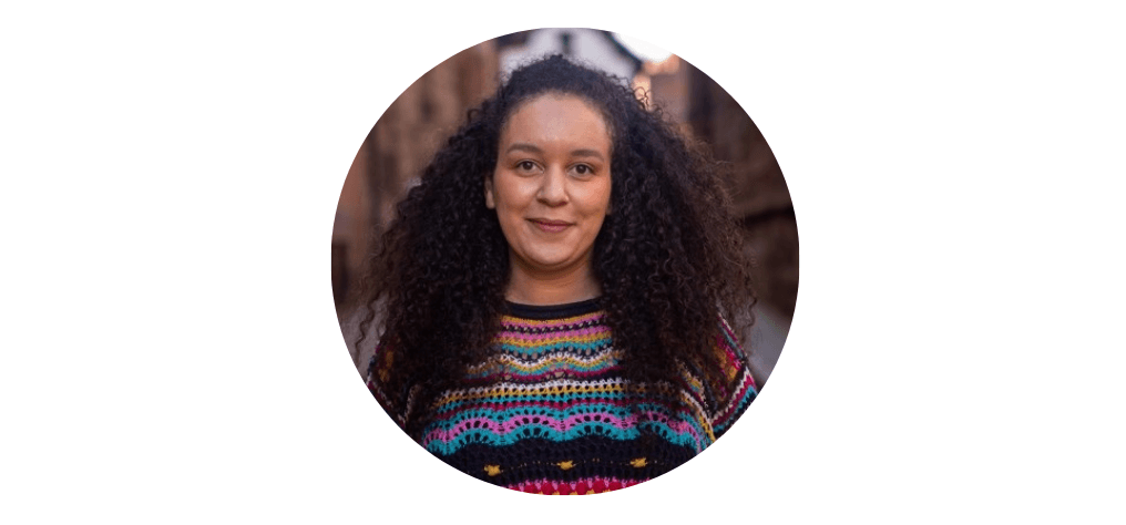 A woman with long curly brown hair. She is looking directly at the camera smiling, wearing a colourful jumper. Her photo is contained with a circular frame.