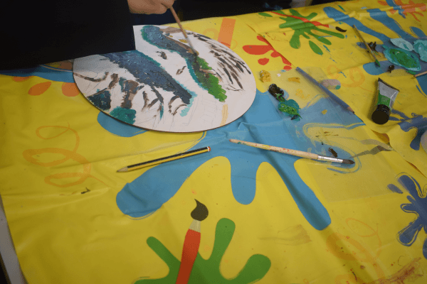 A bright yellow table cover with paint splatter patterns. On top of the cover is paint tubes, paint brushes and a round wooden board being painted.
