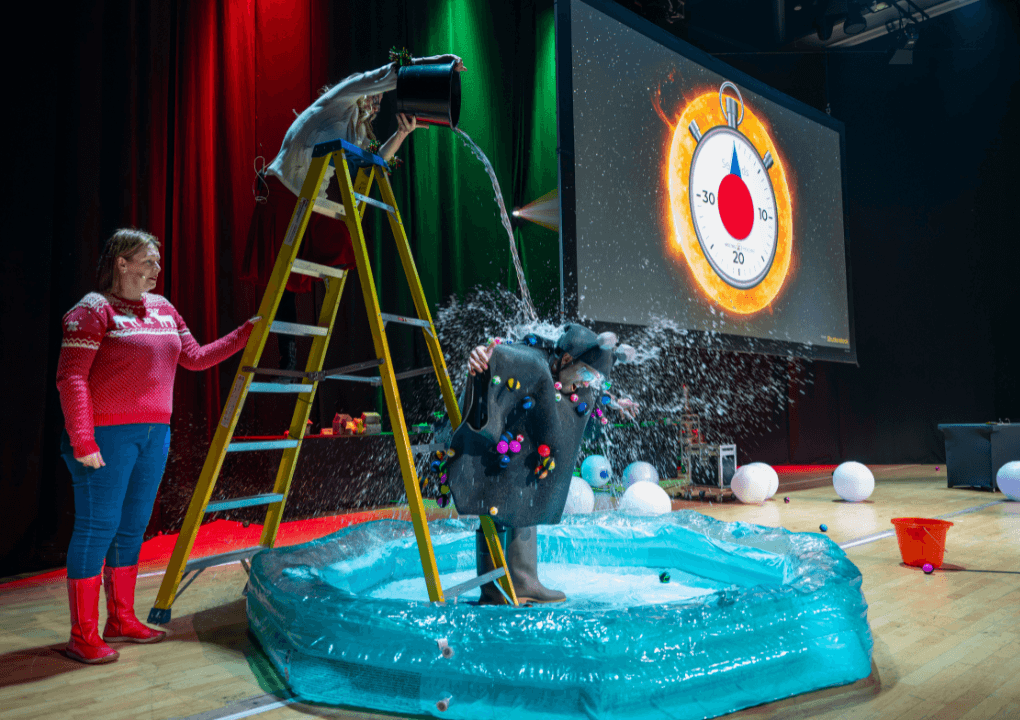 A man dressed as a bug is stood in a blue paddling pool on a stage. Above him is a woman stood on a ladder pouring water over him from a bucket.